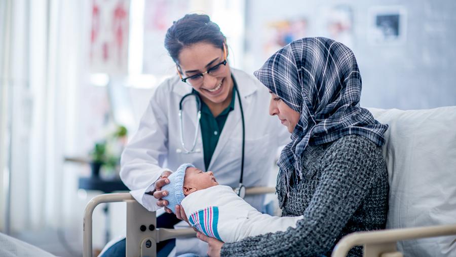 A mother and her newborn baby boy are indoors in a hospital. The mother is holding her baby while laying in the hospital bed. Their doctor is smiling down at the baby.